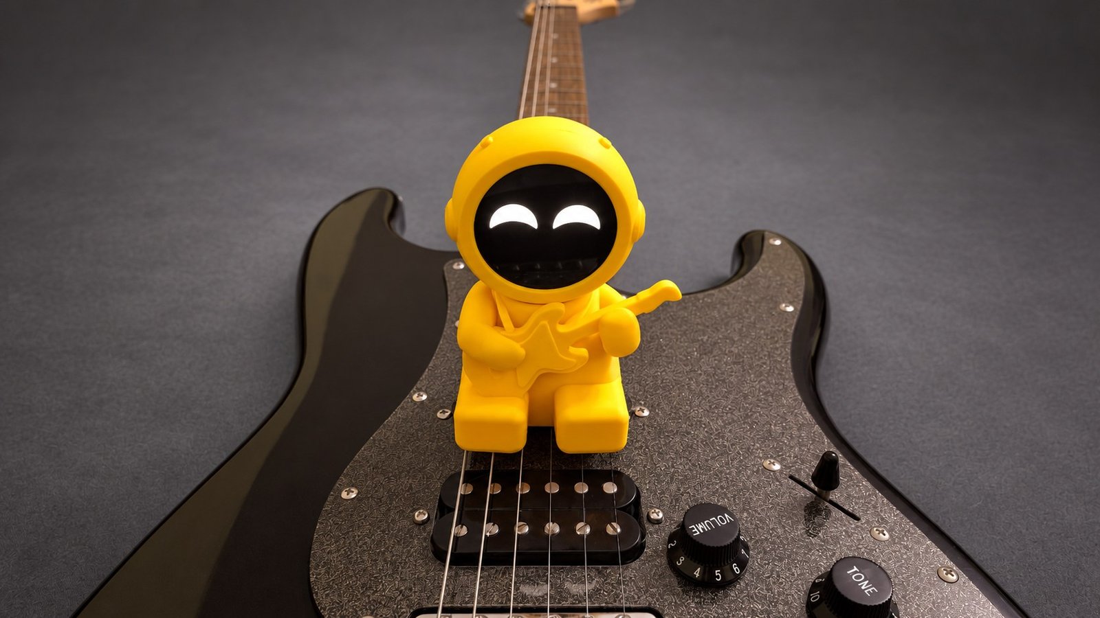 Yellow RW desk robot sitting on a black electric guitar against a charcoal studio background